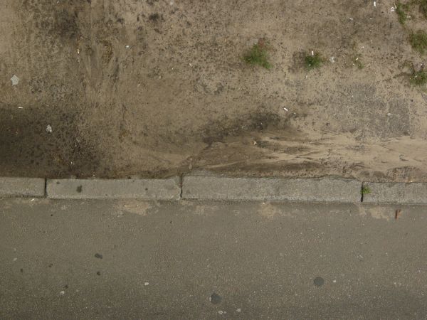 Road edge texture, with a swirling brown dirt surface that has bits of grass and debris, separated by a stone divider from an area of dark road.